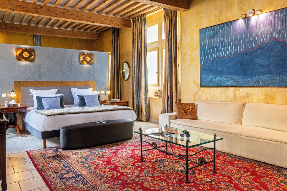 Un dormitorio con cama, mesitas de noche, una mesa de centro de vidrio, un sofá, una alfombra roja estampada y un cuadro grande en la pared.