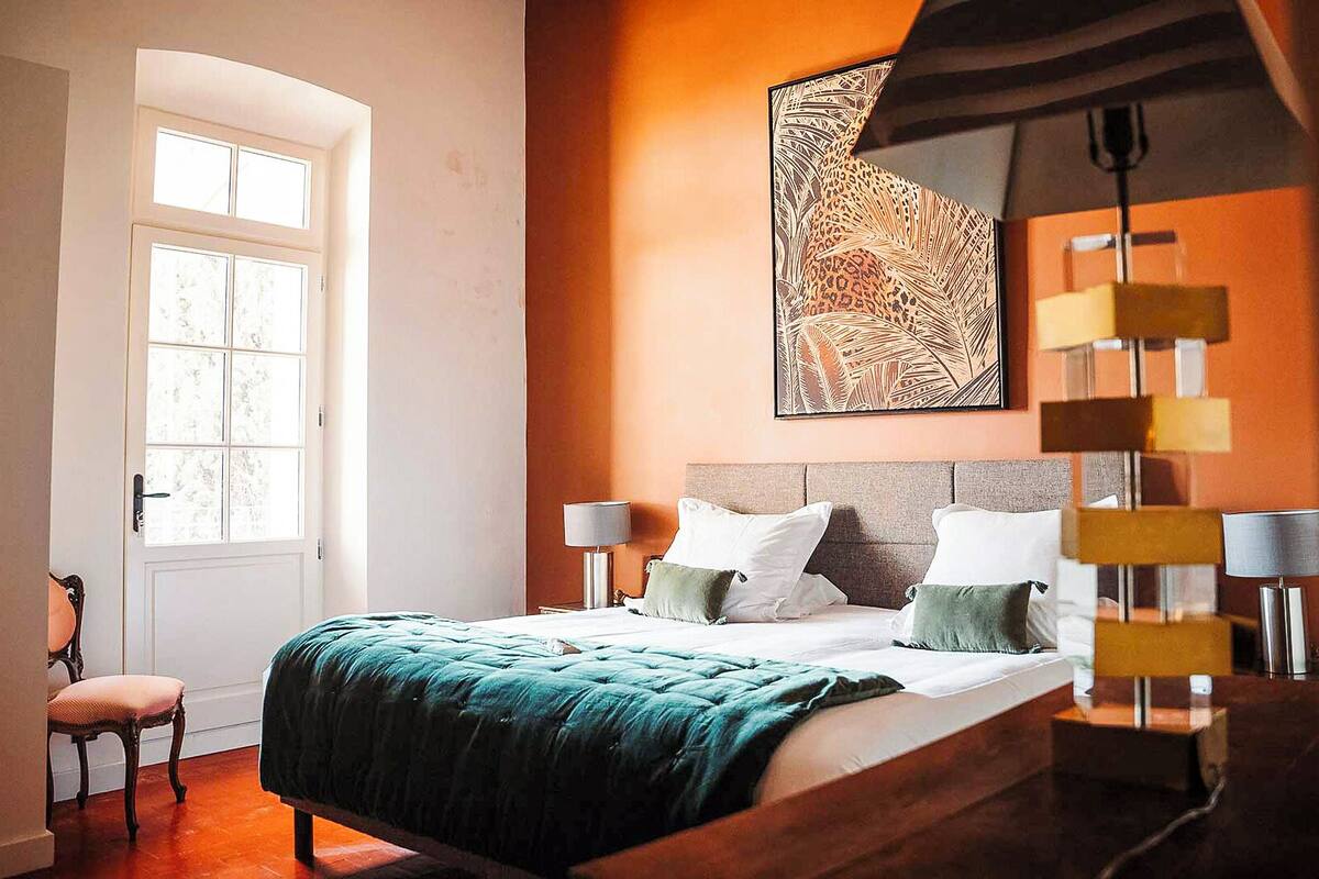 Un dormitorio con una cama, dos mesitas de noche, una silla, un ventanal y una obra de arte colgada en la pared.