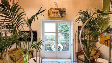 Un invernadero con puerta de cristal que da a un balcón, rodeado de plantas diversas y mobiliario de mimbre.