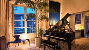 Un piano de cola en una habitación luminosa con ventanas amplias, una planta en maceta, una silla moderna y una lámpara.