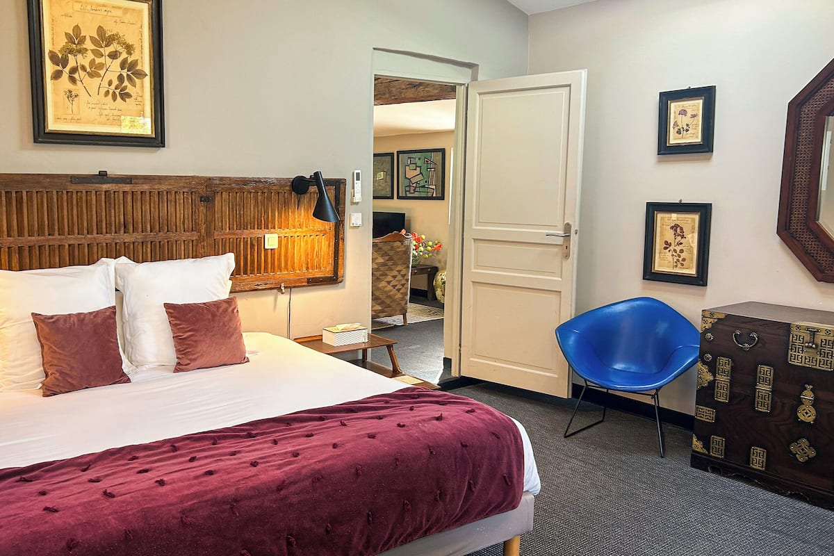 Habitación de hotel con cama, una silla azul, cabecera de madera e impresiones botánicas enmarcadas en las paredes.