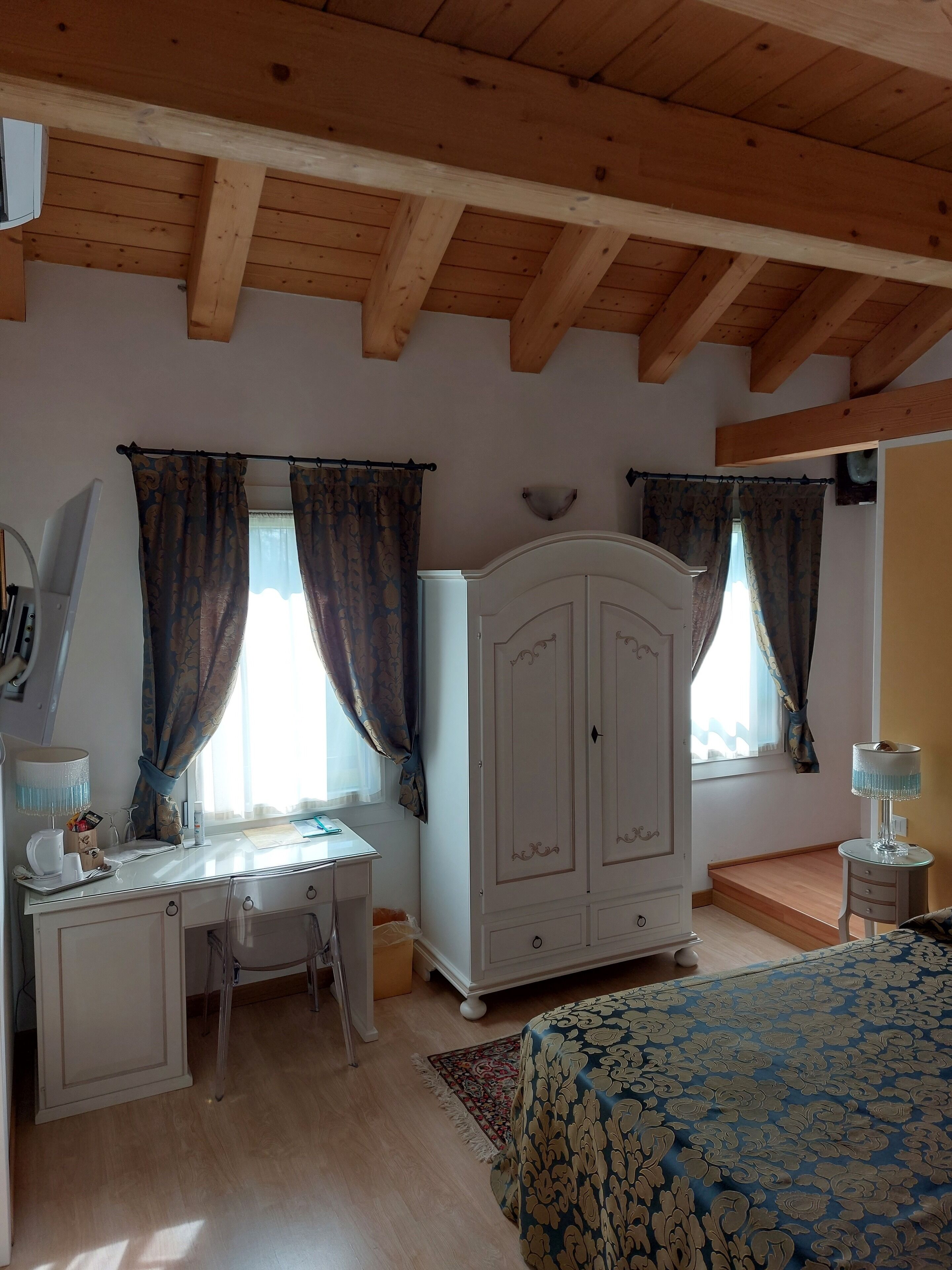 A bedroom with a wooden ceiling, a desk with a lamp, a wardrobe, and a bed with a patterned bedspread.