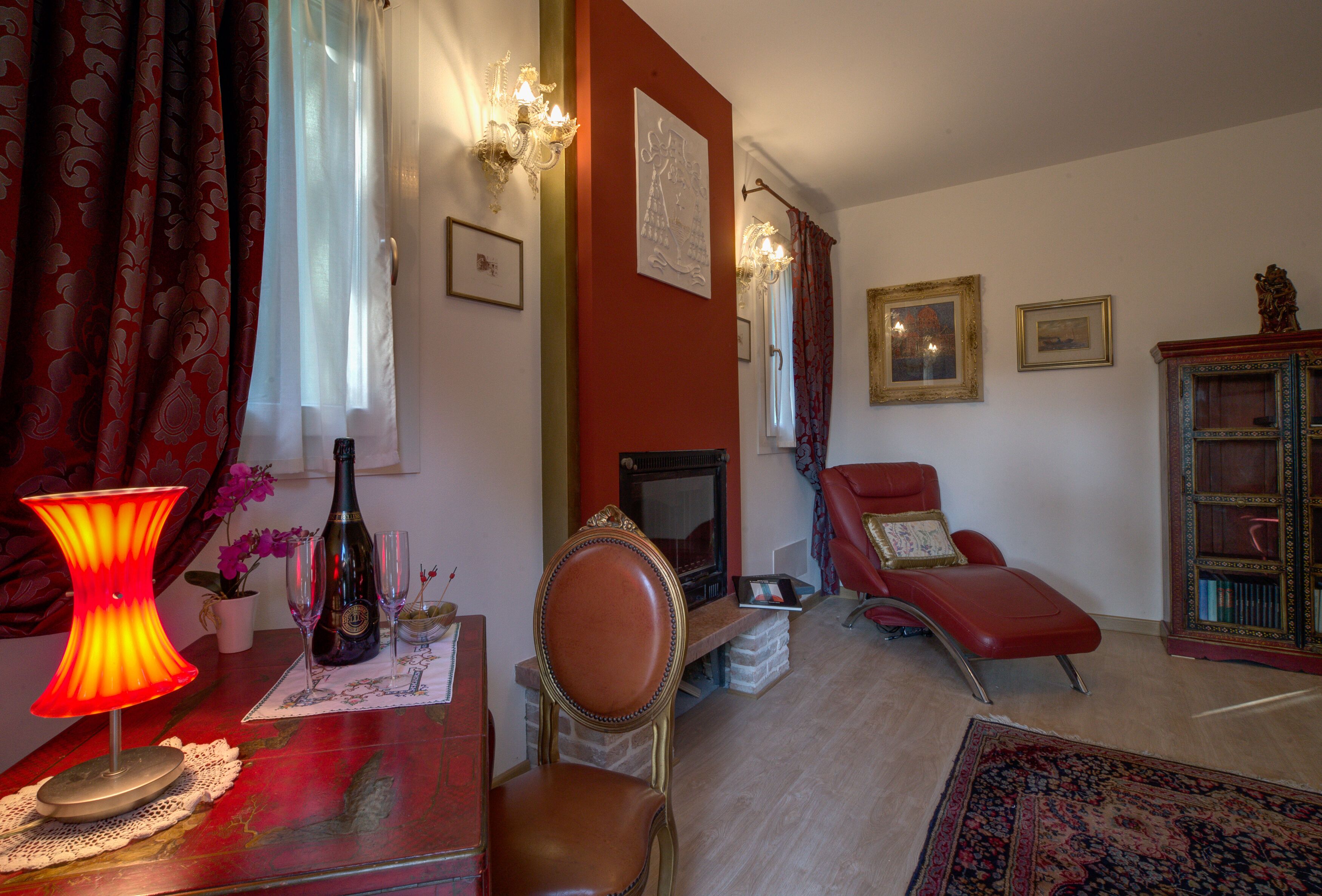 A living room with a fireplace, a red armchair, a red sofa, a red rug, and a table with a lamp and a bottle.