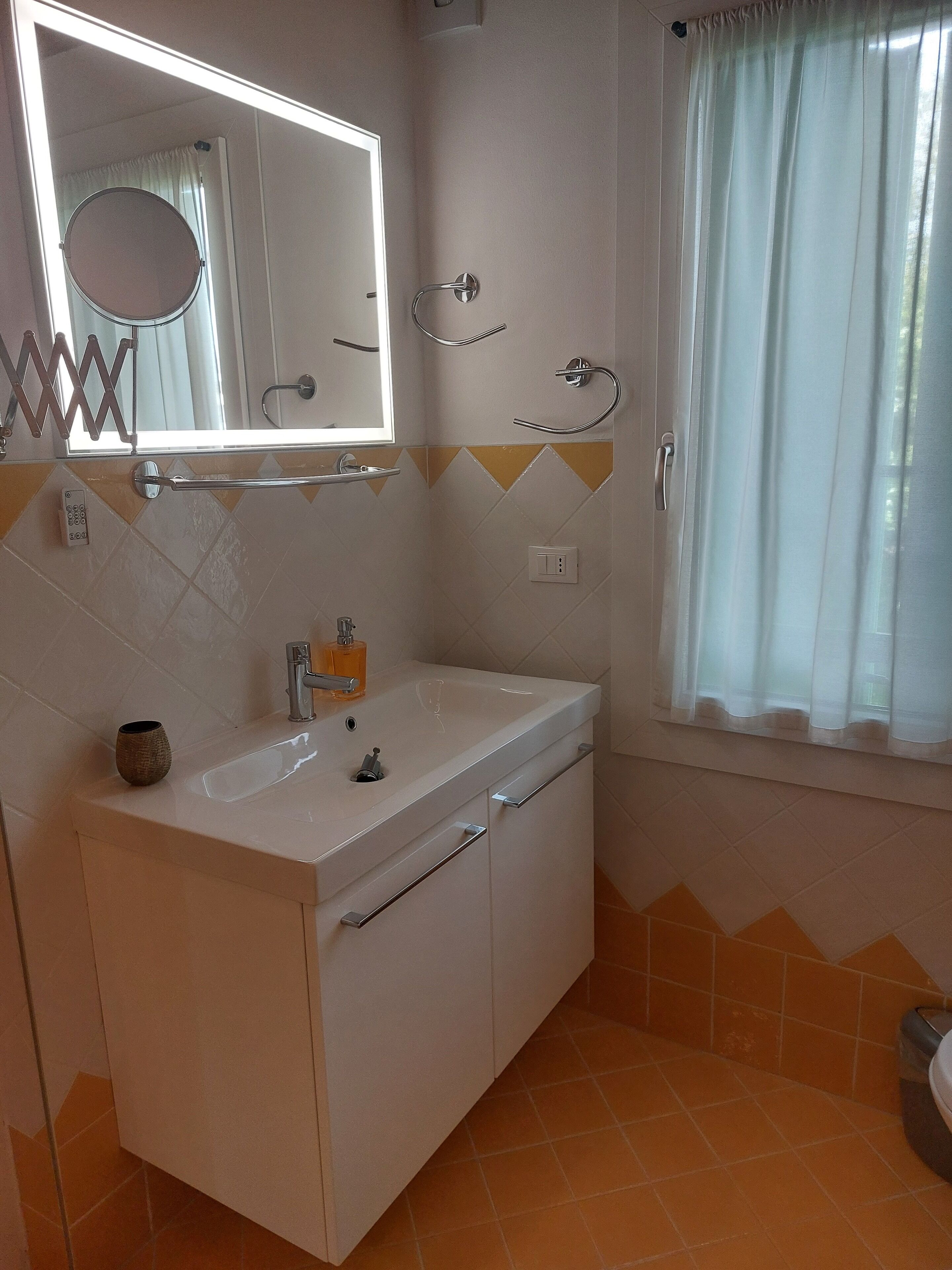 Un baño con lavamanos blanco, espejo y toallero. Se ve una ventana con cortina blanca.