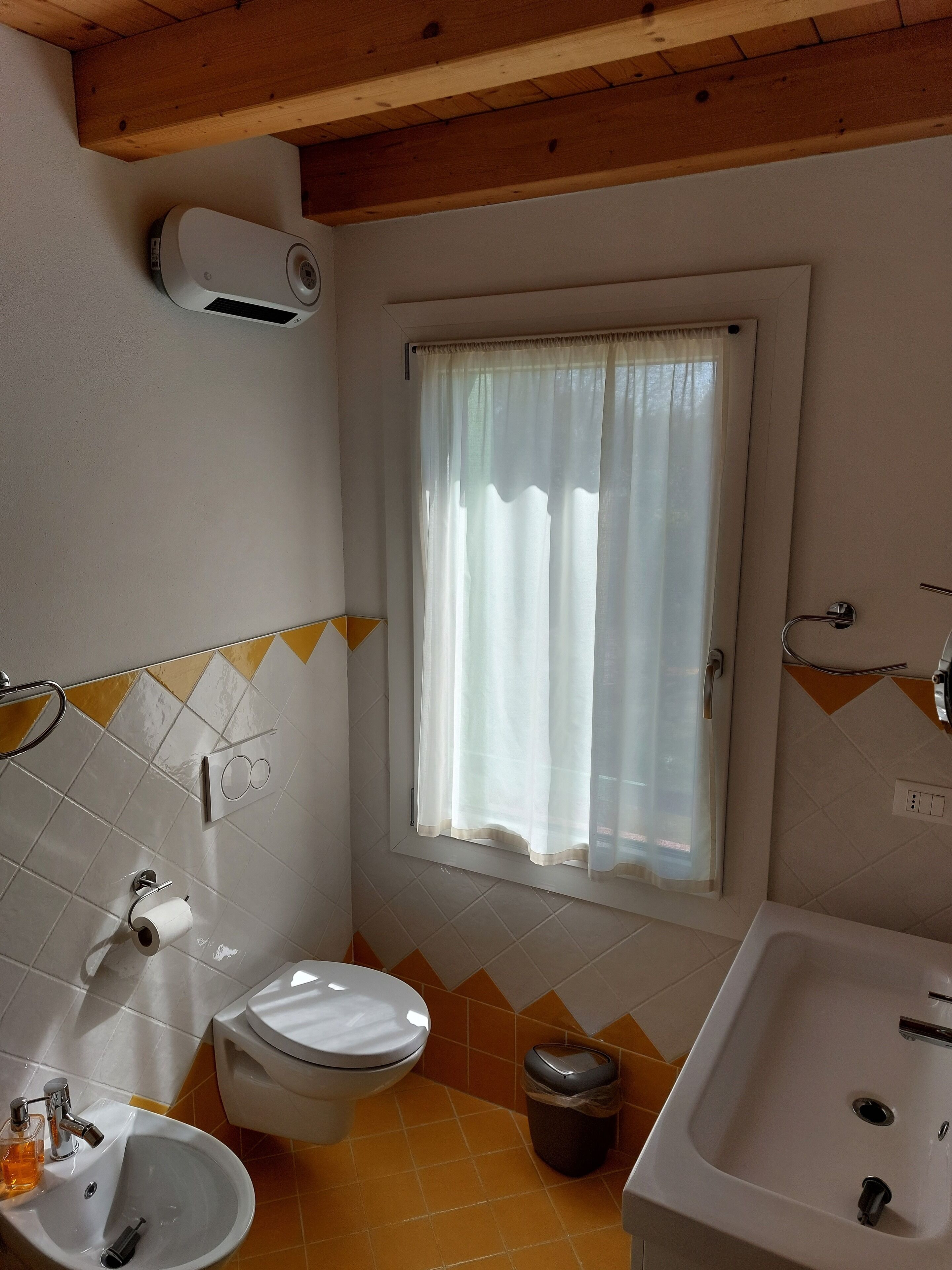 Une salle de bain équipée de toilettes, d’un lavabo et d’une baignoire. Il y a une fenêtre avec un rideau et un climatiseur fixé au mur.