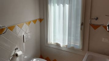 Une salle de bain équipée de toilettes, d’un lavabo et d’une baignoire. Il y a une fenêtre avec un rideau et un climatiseur fixé au mur.
