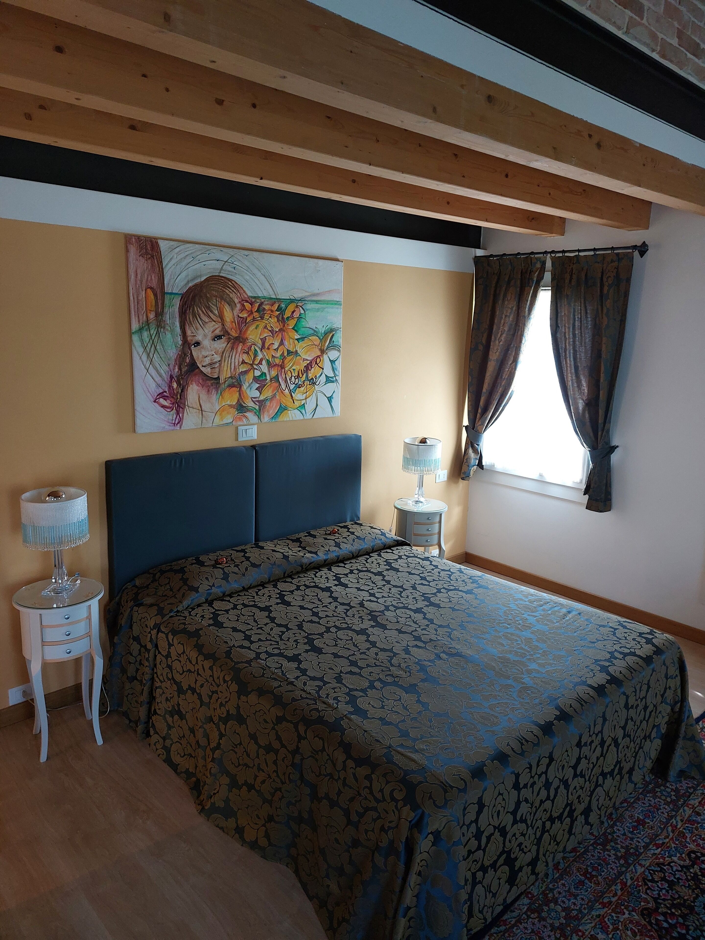 Une chambre à coucher avec un lit, une table de chevet, une lampe, une fenêtre avec des rideaux et un tableau au mur.