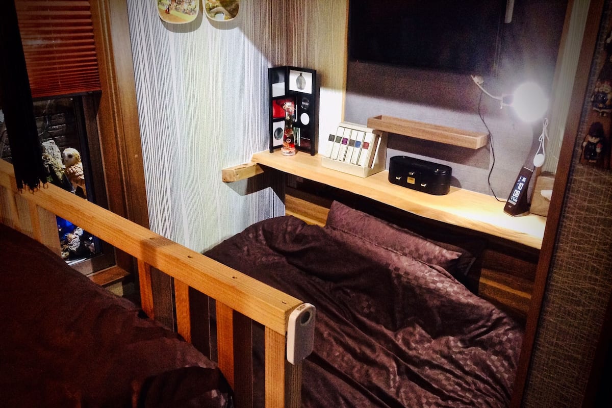 A cozy bedroom with a wooden bed, a nightstand with a lamp, and a shelf with various items.