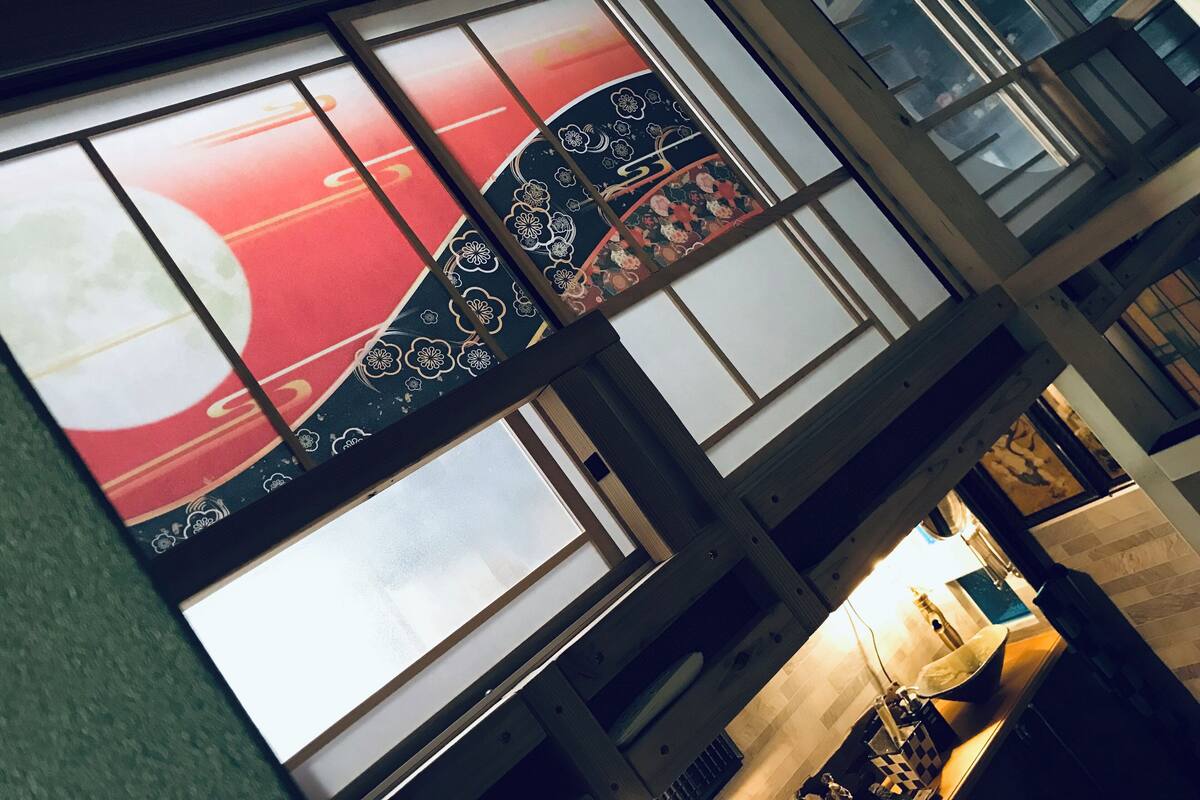 A room with large windows featuring traditional Japanese artwork, a kitchen area with modern appliances, and a view of the cityscape through the windows.