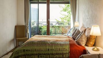 A bedroom with a large bed, a chair, a nightstand with lamps, and a view of a balcony with greenery.