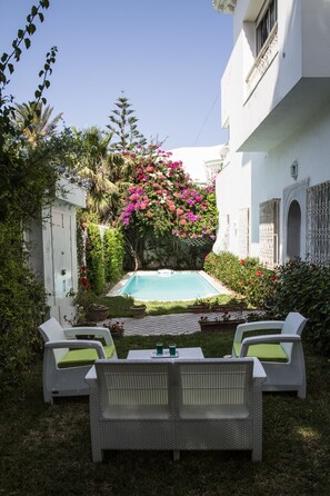 Un área de piscina con paredes blancas, piso a cuadros y dos sillas de mimbre blanco con cojines verdes.