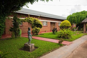 Un edificio de ladrillo con una estatua en primer plano, rodeado de follaje cuidado y un camino pavimentado.