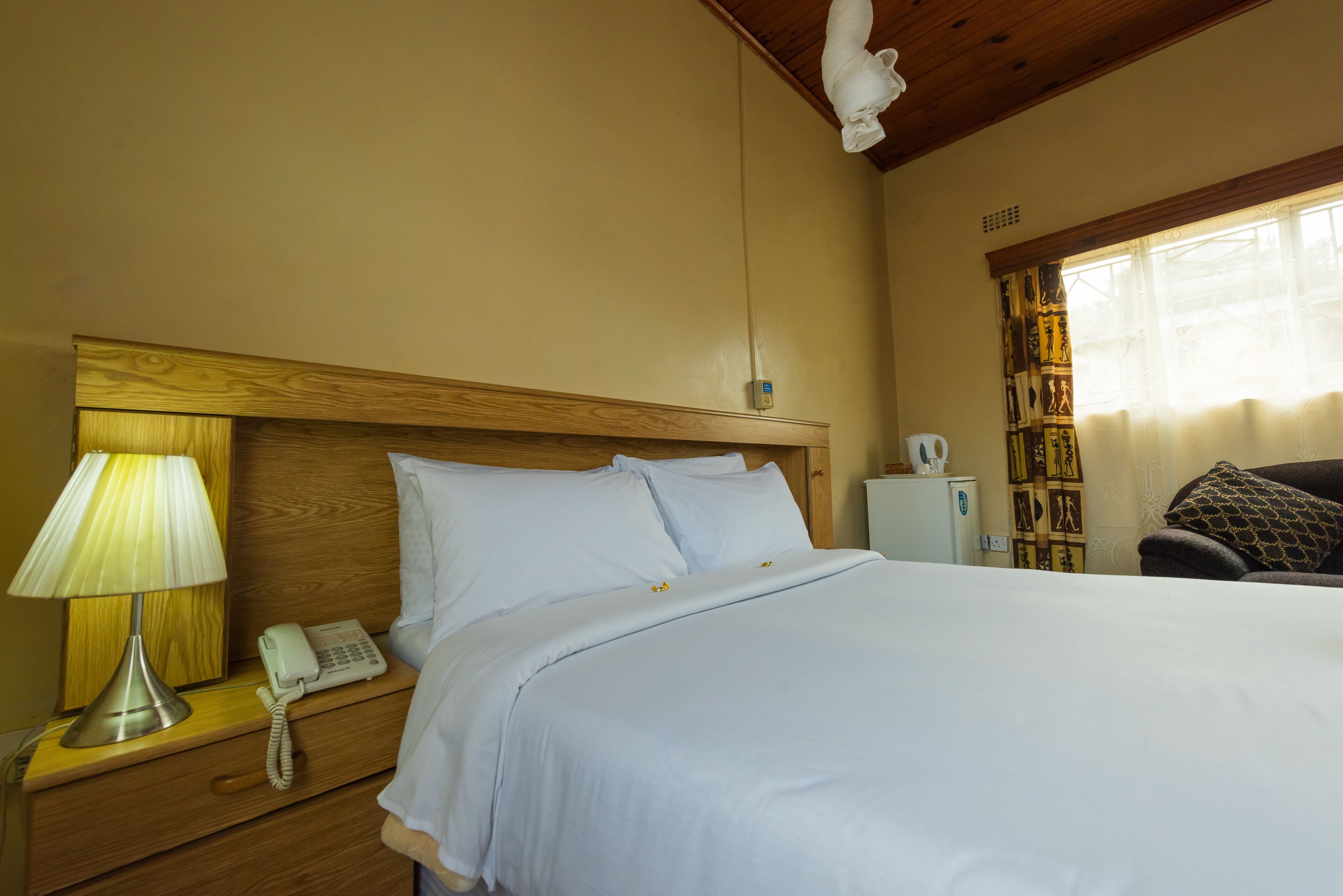 A neatly made bed with white linens, a bedside lamp, a telephone, and a small white appliance on a wooden nightstand.