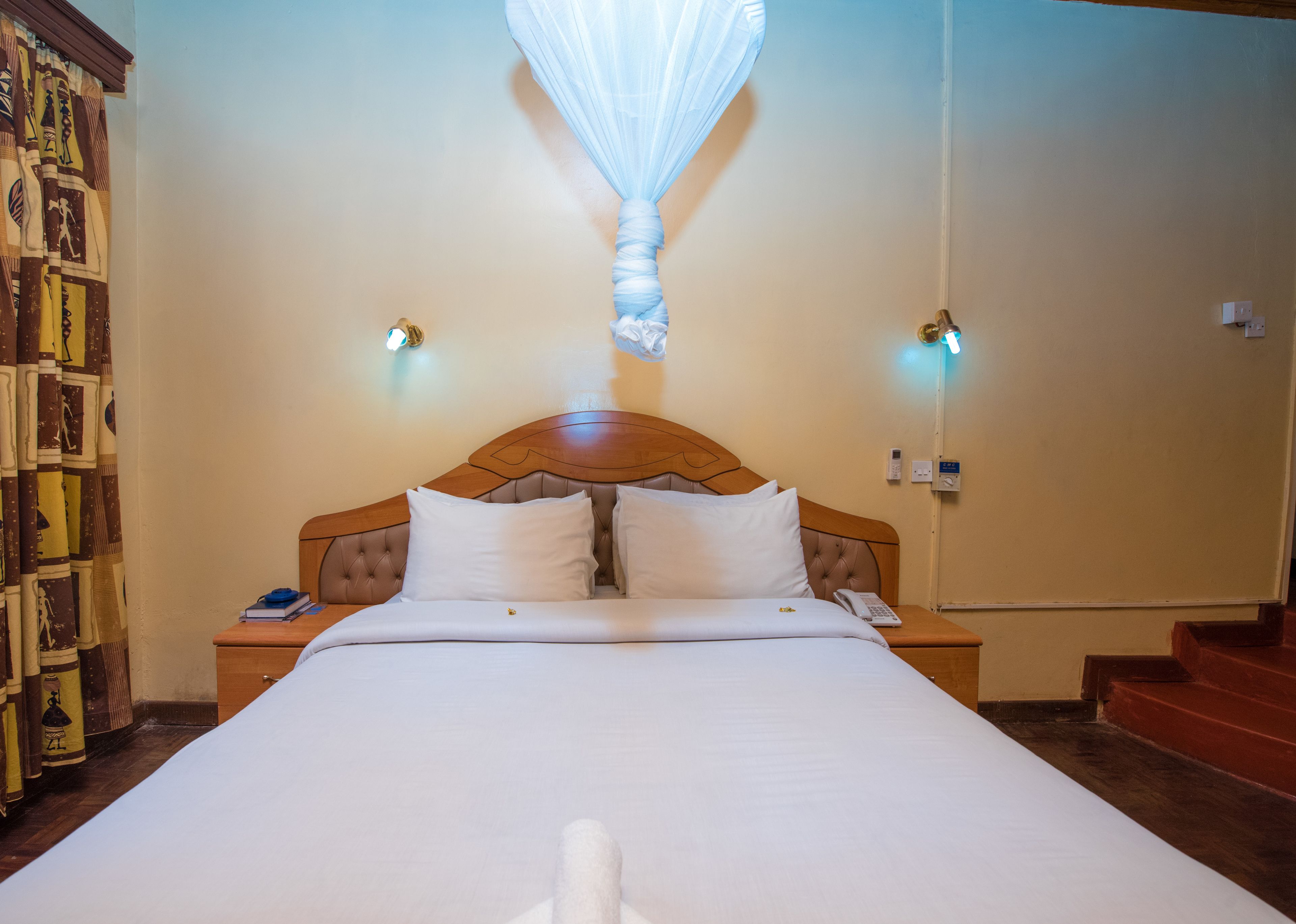 A hotel room with a large bed, two bedside tables, a nightstand, a wall-mounted lamp, and a curtain with Egyptian-themed designs.
