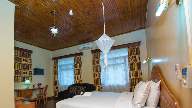 A hotel room with a bed, bedside table, desk, chair, and a wooden ceiling. - Nelly's Guesthouse (Lilongwe)