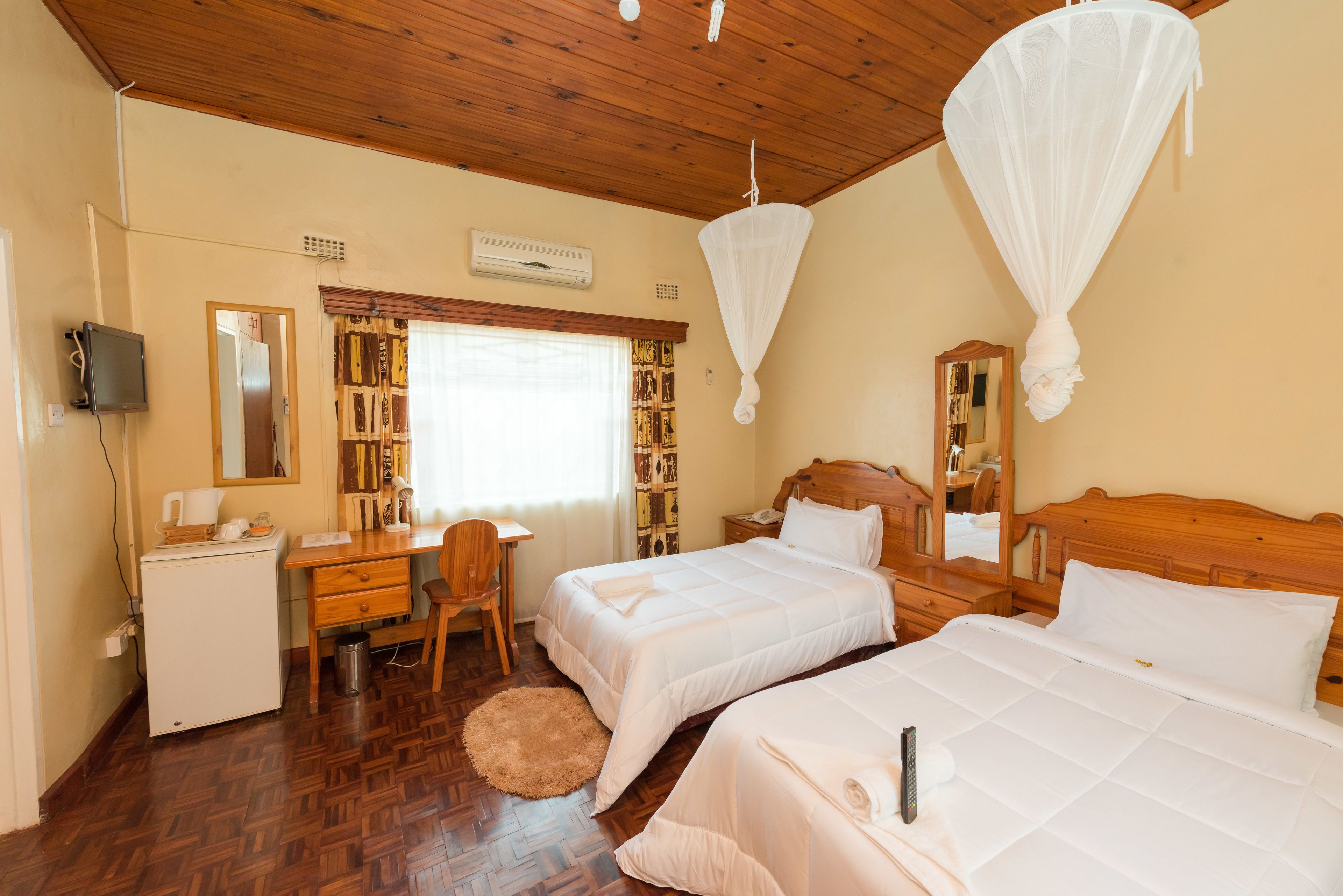 A hotel room with two beds, a desk, a chair, a small refrigerator, and a bookshelf.