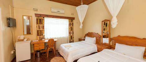 A hotel room with two beds, a desk, a chair, a small refrigerator, and a bookshelf.
