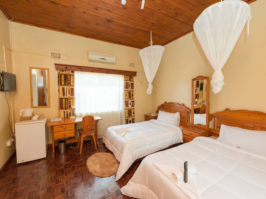A hotel room with two beds, a desk, a chair, a small refrigerator, and a bookshelf.
