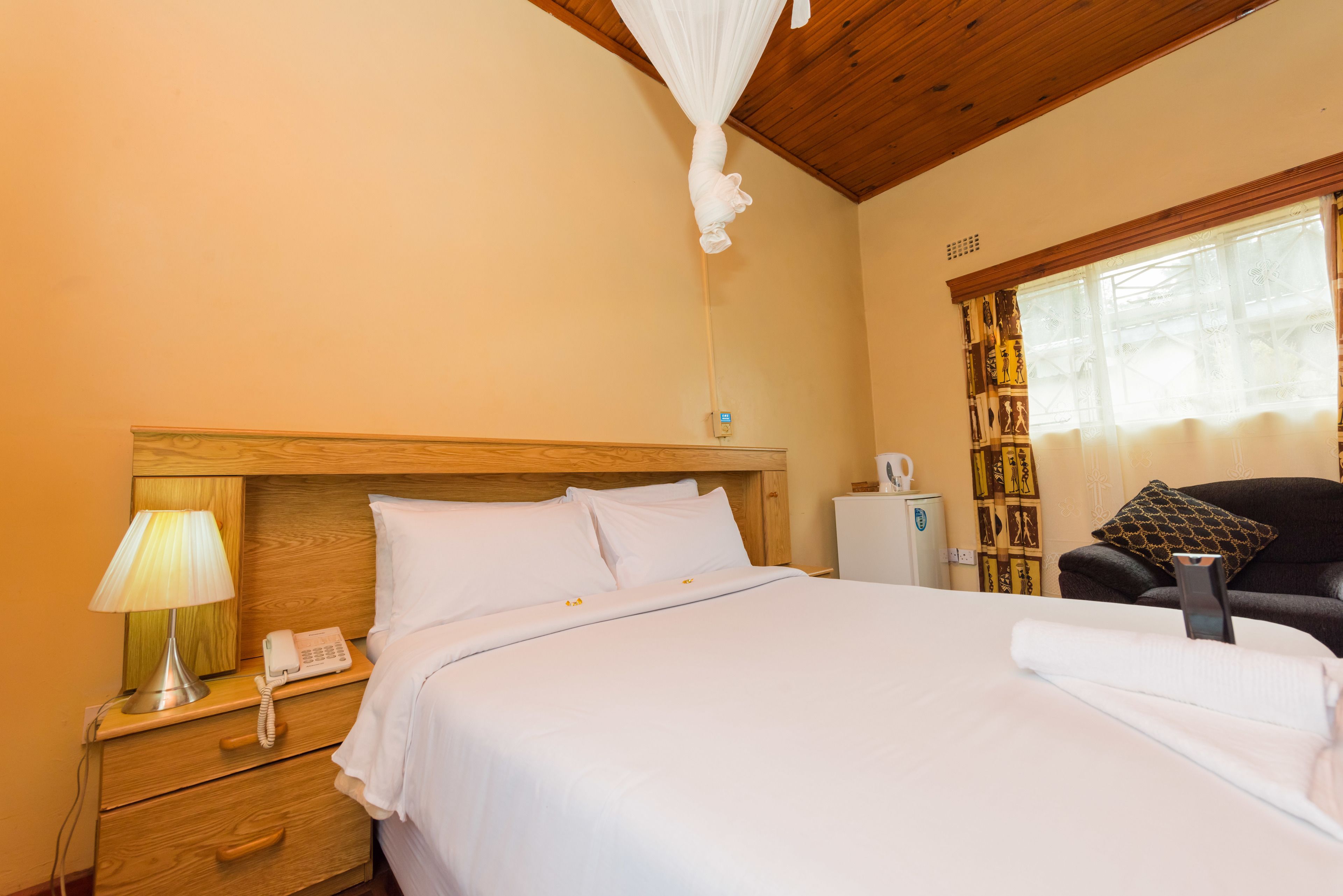 Habitación de hotel con cama, mesita de noche con lámpara, teléfono, silla y ventana con cortinas.