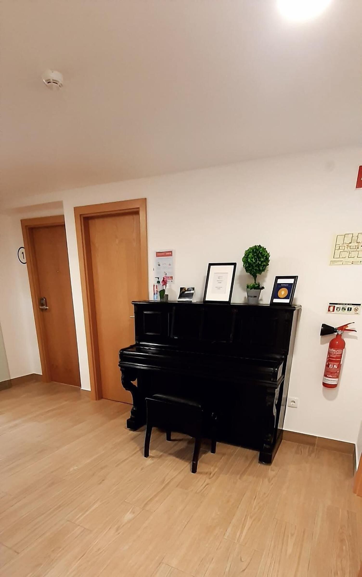 A room with a black piano, wooden flooring, two wooden doors, a fire extinguisher, and a small potted plant on the wall.