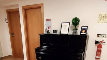 A room with a black piano, wooden flooring, two wooden doors, a fire extinguisher, and a small potted plant on the wall.