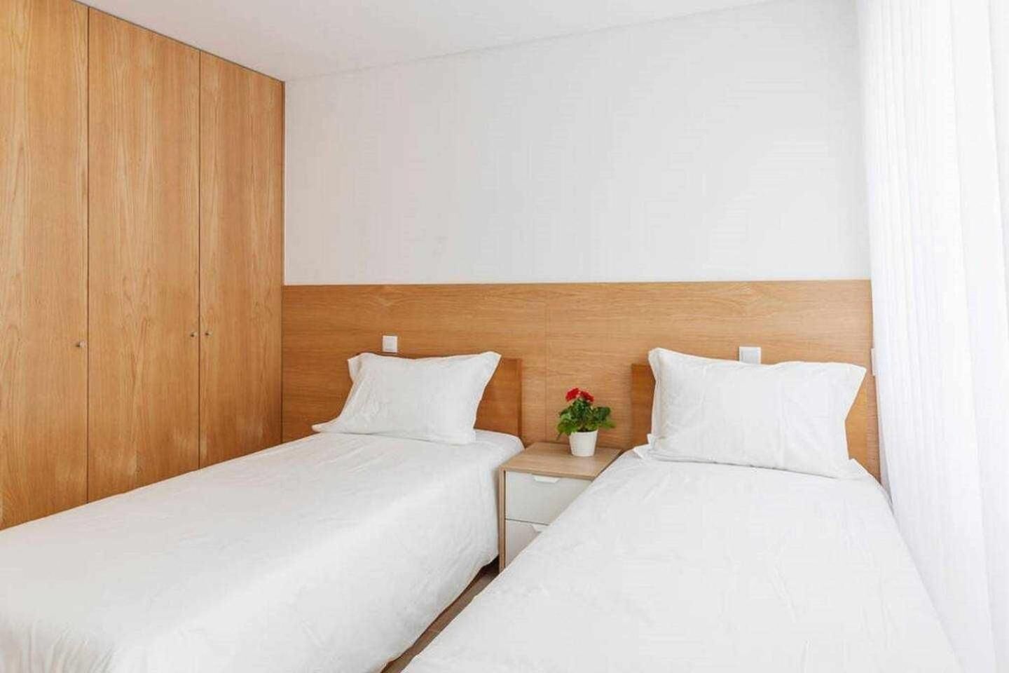 Een kamer met twee eenpersoonsbedden, houten hoofdeinden, wit beddengoed en een klein nachtkastje met een rode bloem.