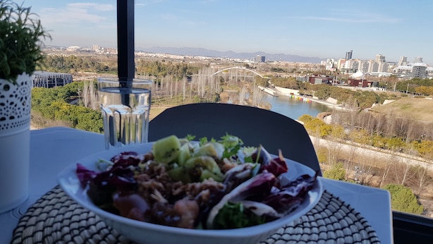 Une table avec un saladier rempli de salade et un verre d’eau, donnant sur un paysage urbain avec des bâtiments et une rivière.