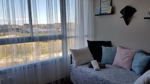 Une chambre avec un lit, une fenêtre ornée de rideaux légers, une étagère fixée au mur avec des photos, et une vue sur la ville.