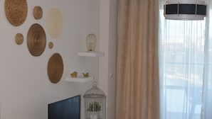 Un salon moderne avec un téléviseur à écran plat posé sur un meuble blanc, un parquet, une table à manger et une grande fenêtre ornée de rideaux légers.