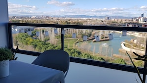 Une pièce dotée d’une grande fenêtre donnant sur un paysage urbain, une table avec un pot de fleurs et une chaise.