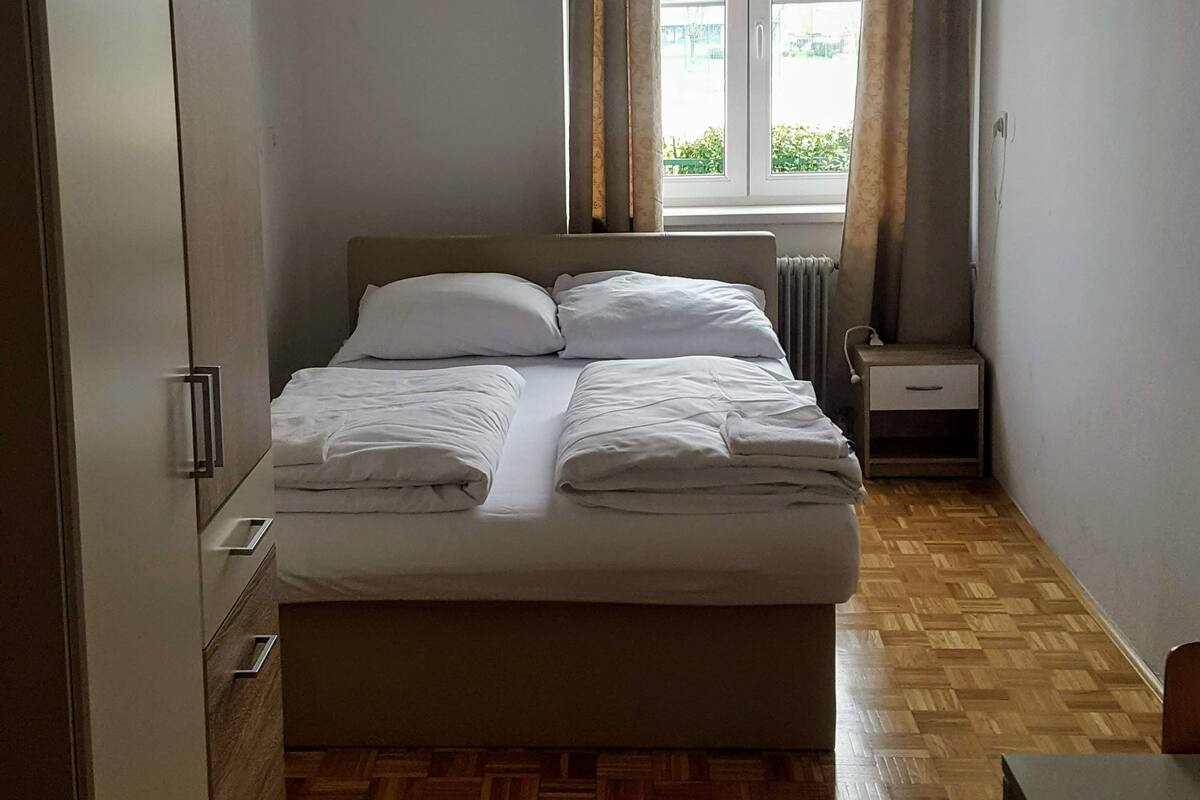 Ein ordentlich bezogenes Bett mit weißen Leinen, Holzboden, ein Nachttisch mit Lampe und ein Fenster mit Vorhängen.