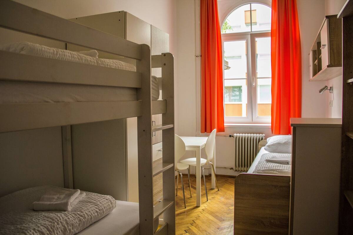 Ein Schlafraum mit Etagenbetten, Schreibtisch, Stuhl, Heizkörper und einem Fenster mit Vorhängen.