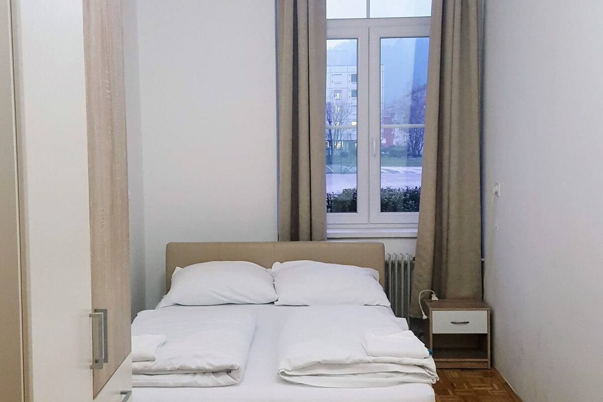 Ein ordentlich bezogenes Bett in einem Zimmer mit großem Fenster, Holzdielenboden und Heizkörper.