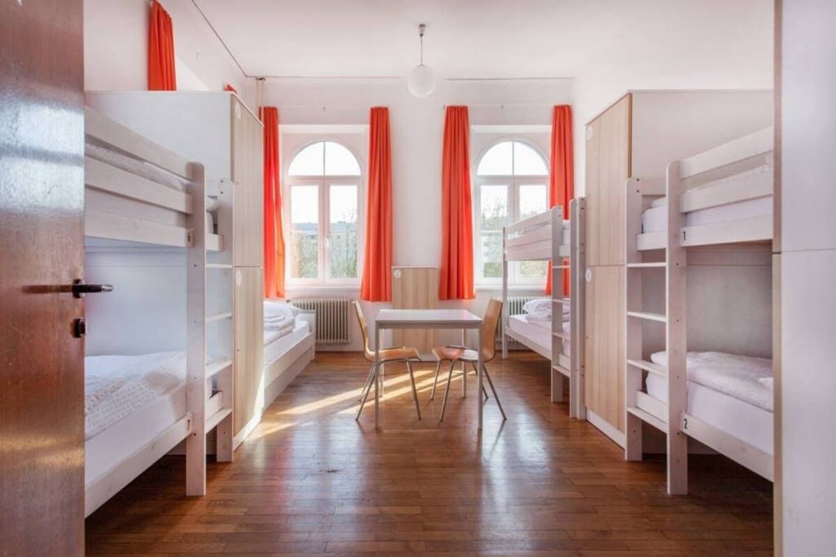 Ein Schlafsaalzimmer mit Etagenbetten, einem Tisch, Stühlen und großen Fenstern mit Vorhängen.
