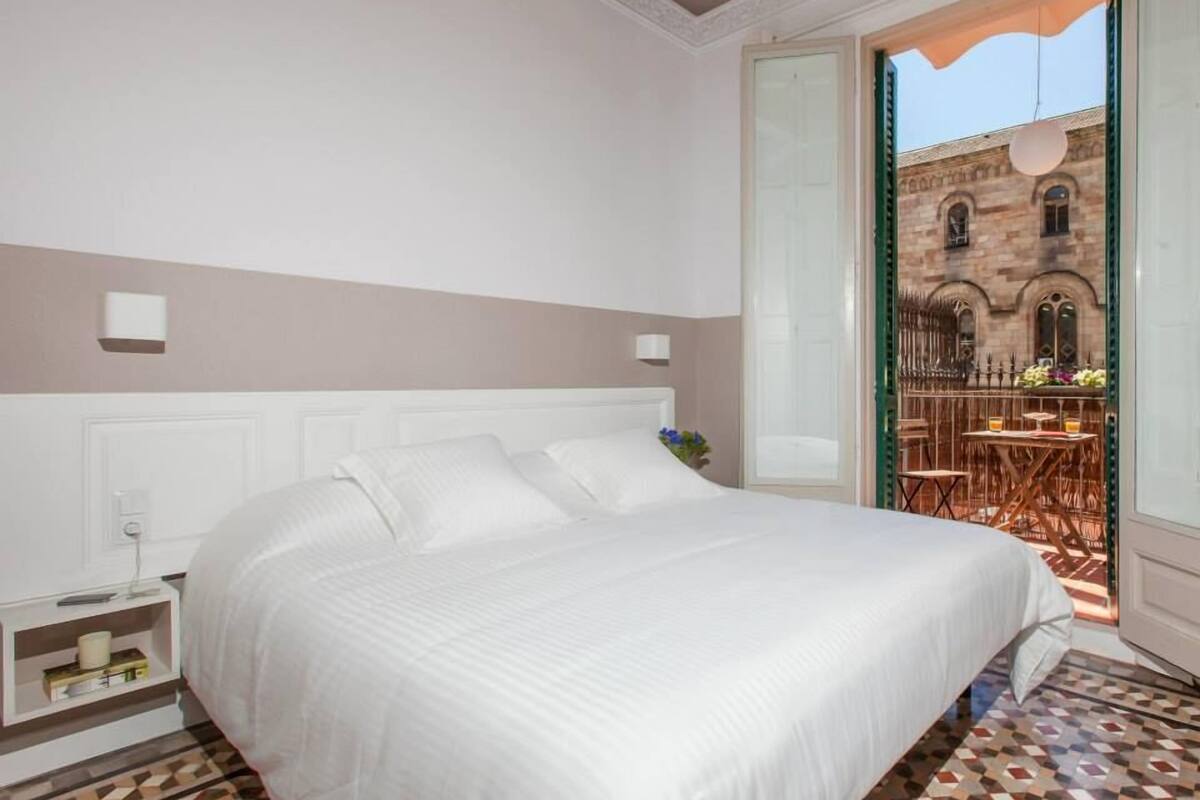 Una habitación de hotel con una cama grande, una mesita de noche, una lámpara de pared y vistas a un patio exterior con mesas y sillas.