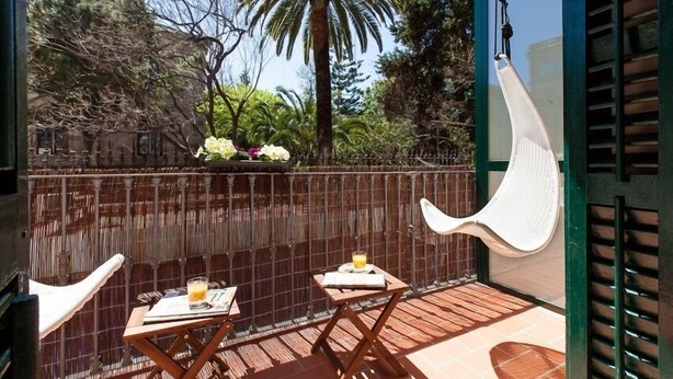 A balcony with two wooden tables, a hanging chair, and a view of trees and a palm tree. - Ecozentric (Barcelona)