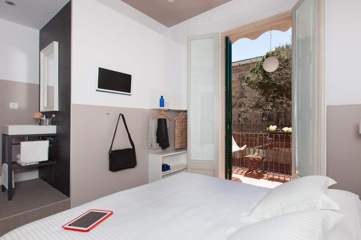 Habitación de hotel moderna con cama, mesita de noche, televisión, baño con lavamanos y espejo, y balcón con vistas al exterior.