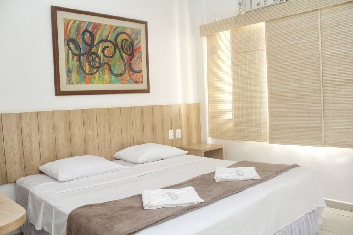 Ein Hotelzimmer mit einem hölzernen Kopfteil, einem Bett mit weißen Leinen, einer braunen Tagesdecke und einem farbenfrohen abstrakten Gemälde an der Wand.