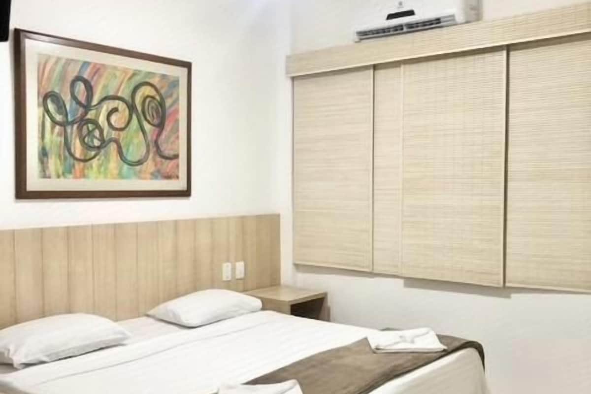 Una habitación de hotel con cama, mesita de noche, aire acondicionado de pared y un cuadro.