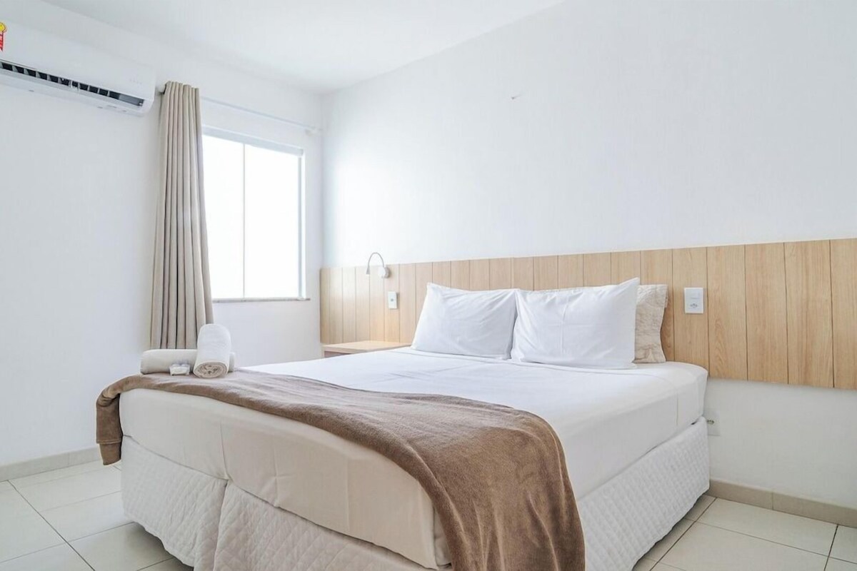 A hotel room with a large bed, a wooden headboard, a window with curtains, and a wall-mounted air conditioner.