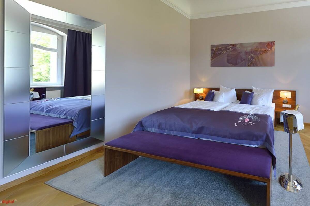 A modern bedroom with a large mirror, a bed with a purple bedspread, bedside lamps, and a painting on the wall.