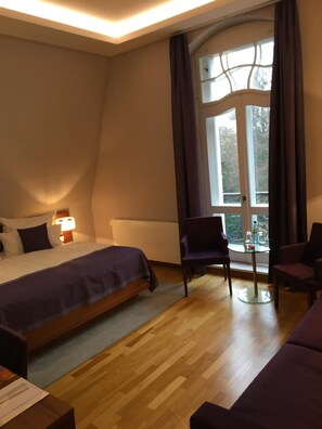 A hotel room with a bed, a small table with chairs, a window with curtains, and a view of trees outside.
