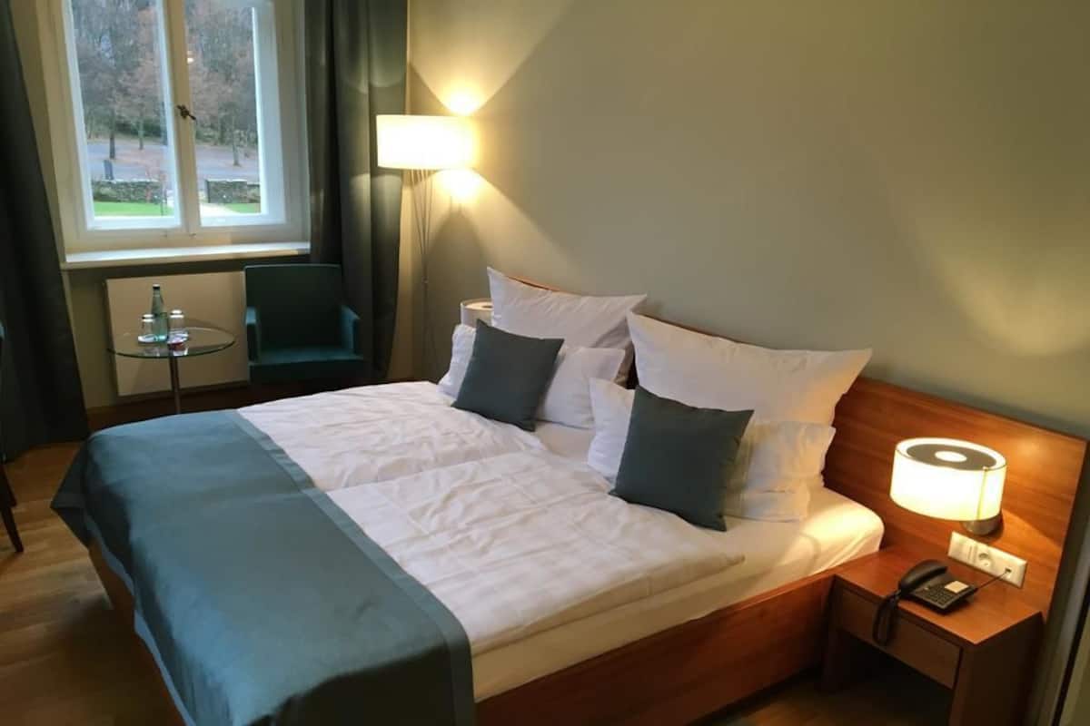 A neatly made bed with white and dark blue bedding, a bedside lamp, a small table with a chair, and a window with a view of trees.