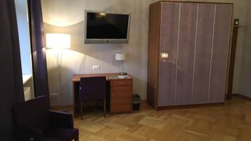 A room with a wooden desk, a chair, a television mounted on the wall, a wardrobe, and a glass table with a bottle and a tissue box.
