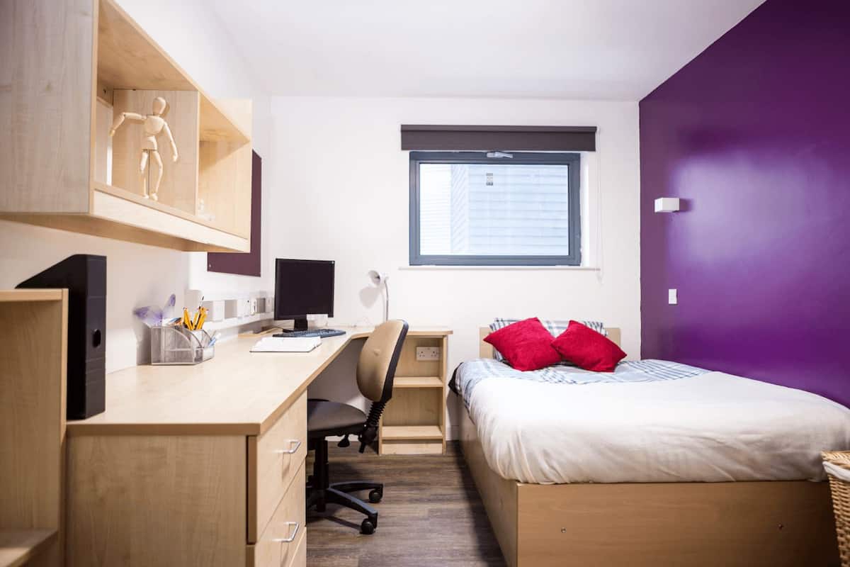 Quarto com escrivaninha de madeira, computador, cadeira, cama com almofadas vermelhas e janela com persianas.