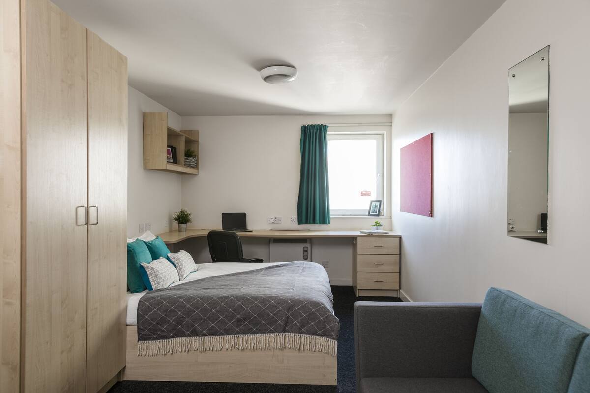 Quarto de hotel com cama, escrivaninha, cadeira, guarda-roupa e janela com cortinas.