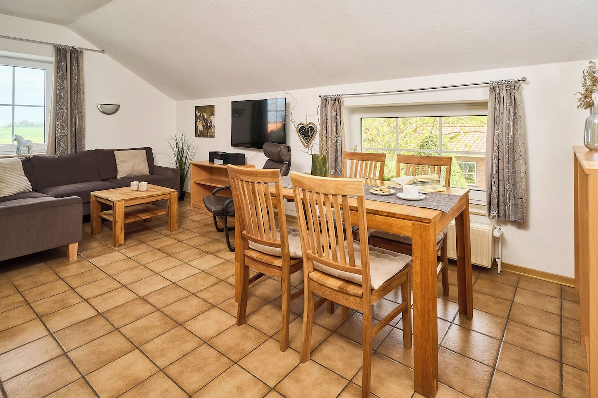 A cozy living room with a sofa, wooden dining table, and chairs. A flat-screen TV is mounted on the wall, and there is a window with curtains.