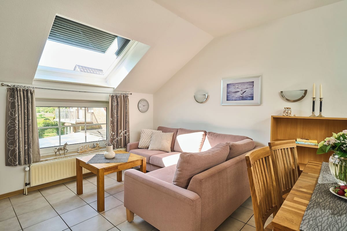 A cozy living room with a sloped ceiling, a sofa, a wooden table, and a view through a large window.