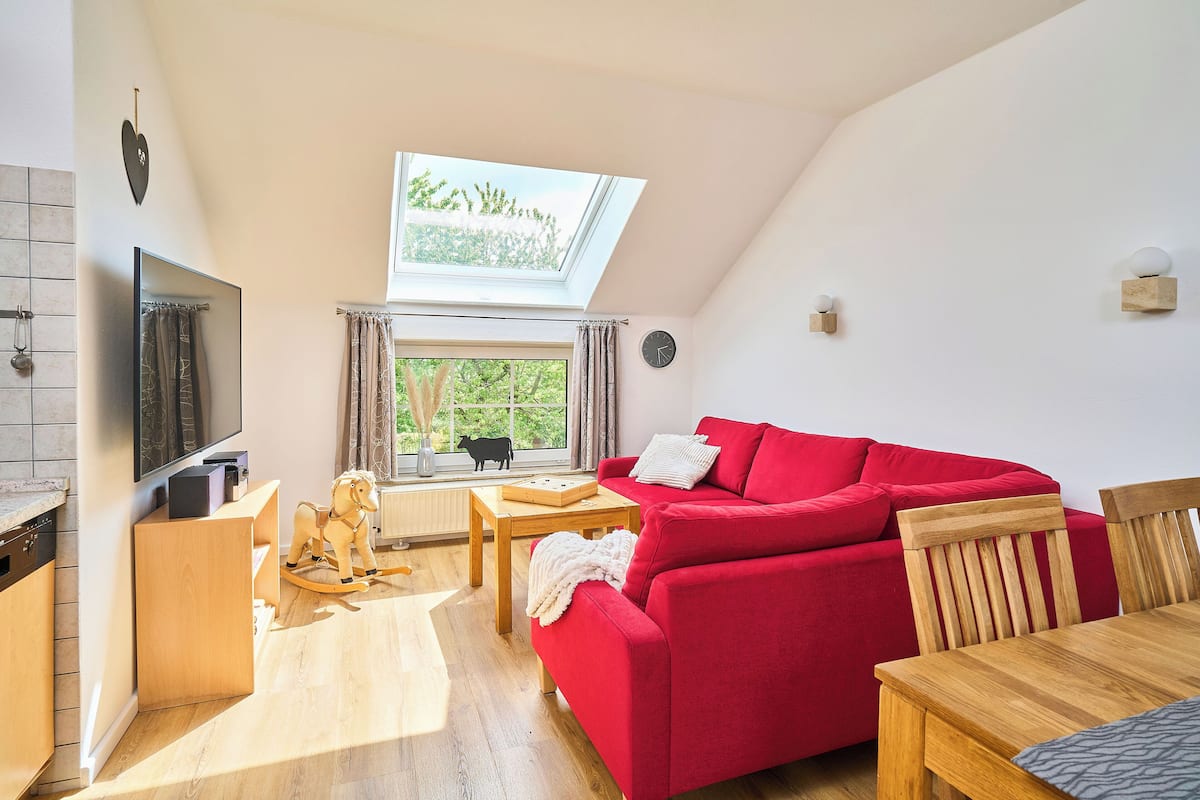 A modern living room with a red sofa, wooden furniture, a TV, and a window with a view of greenery.