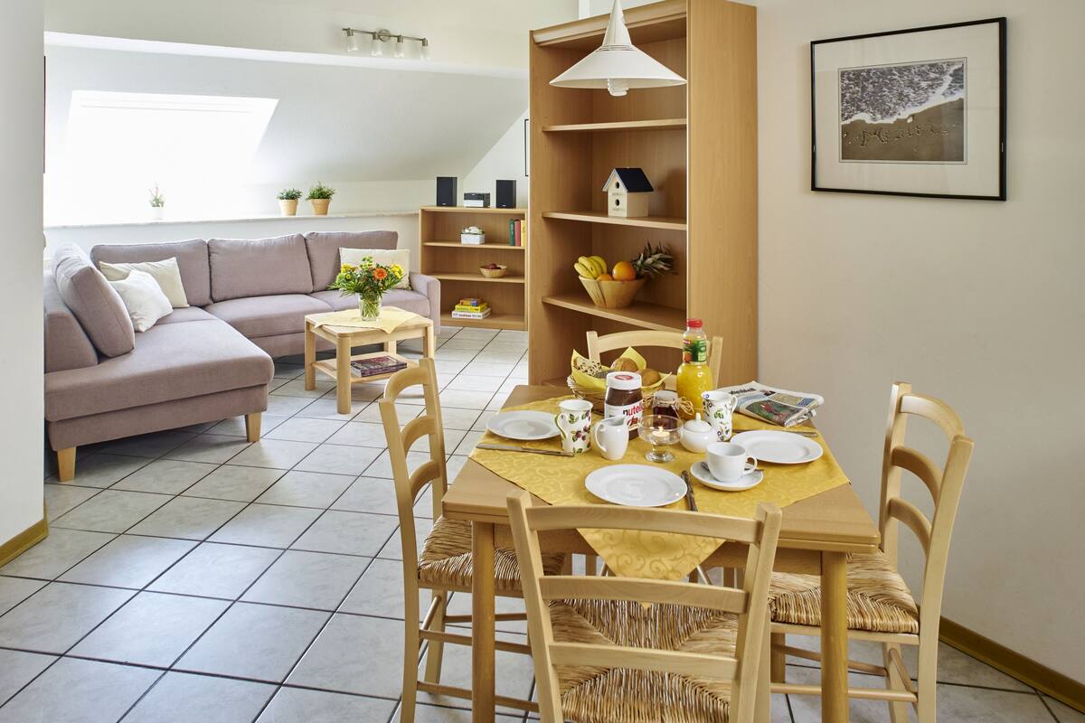 A cozy living room with a dining area, a sofa, a bookshelf, and a dining table set with plates and cups.
